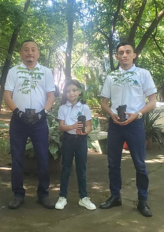 Dos adultos y una niña con camisetas institucionales de Gold Metals, cada uno sosteniendo una planta en maceta, en un área arbolada.