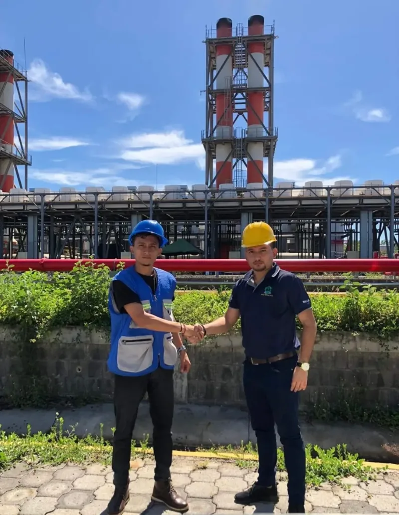 Dos personas en zona industrial con estructura metálica al fondo.