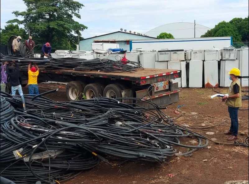 Acopio de rollos de cable junto a camión de plataforma, con personal supervisando la operación y electrodomésticos apilados al fondo.
